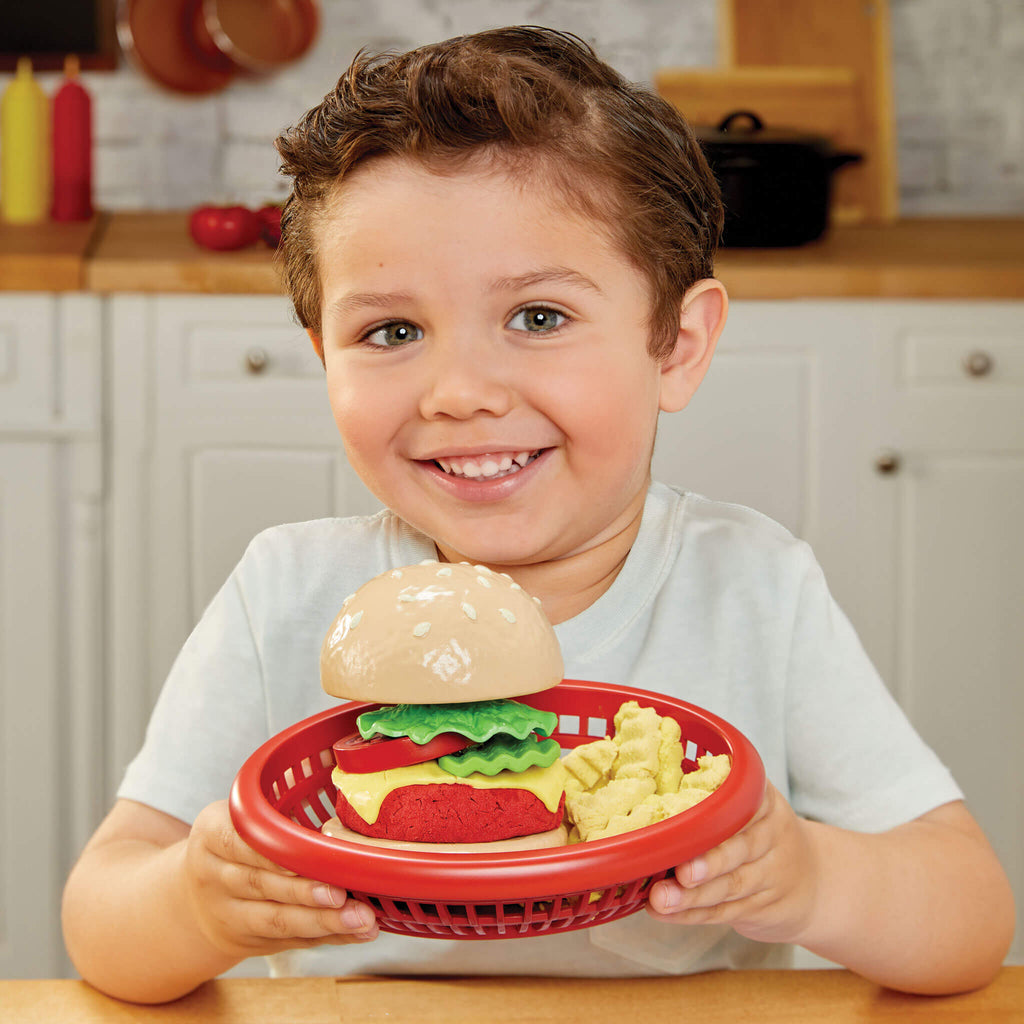 Child holding a toy hamburger set in a red basket in a kitchen setting